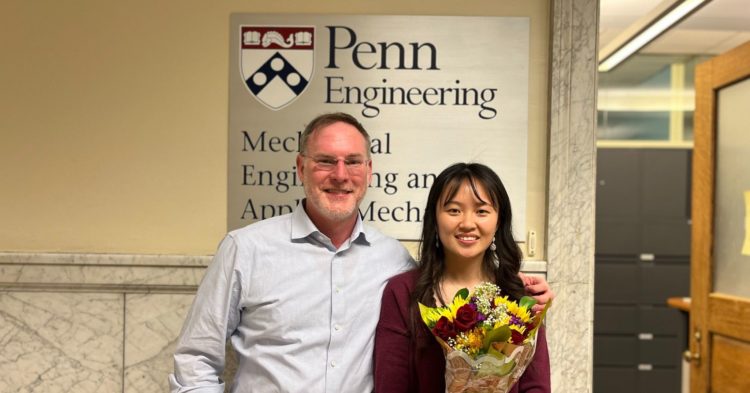 Professor Rob Carpick and Doctor Lu Fang stand in front of the Penn Mechanical Engineering and Applied Mechanics offices. Lu is wearing a maroon shirt and is holding a bouquet of maroon and yellow flowers. 